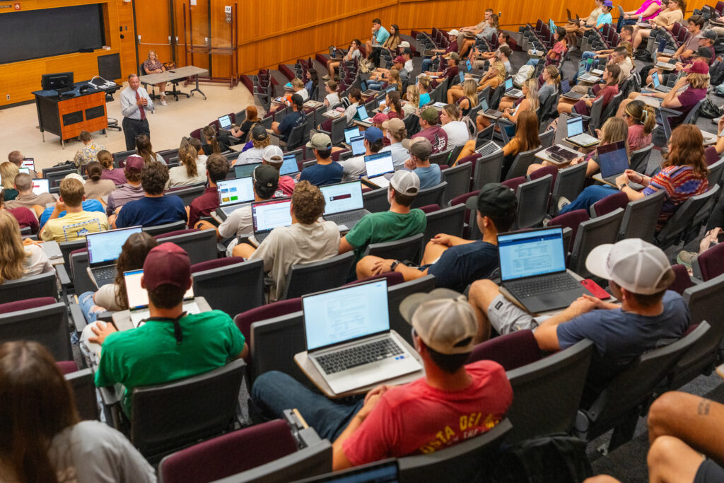 Students sitting in a large lecture hall with laptops open in front of them.