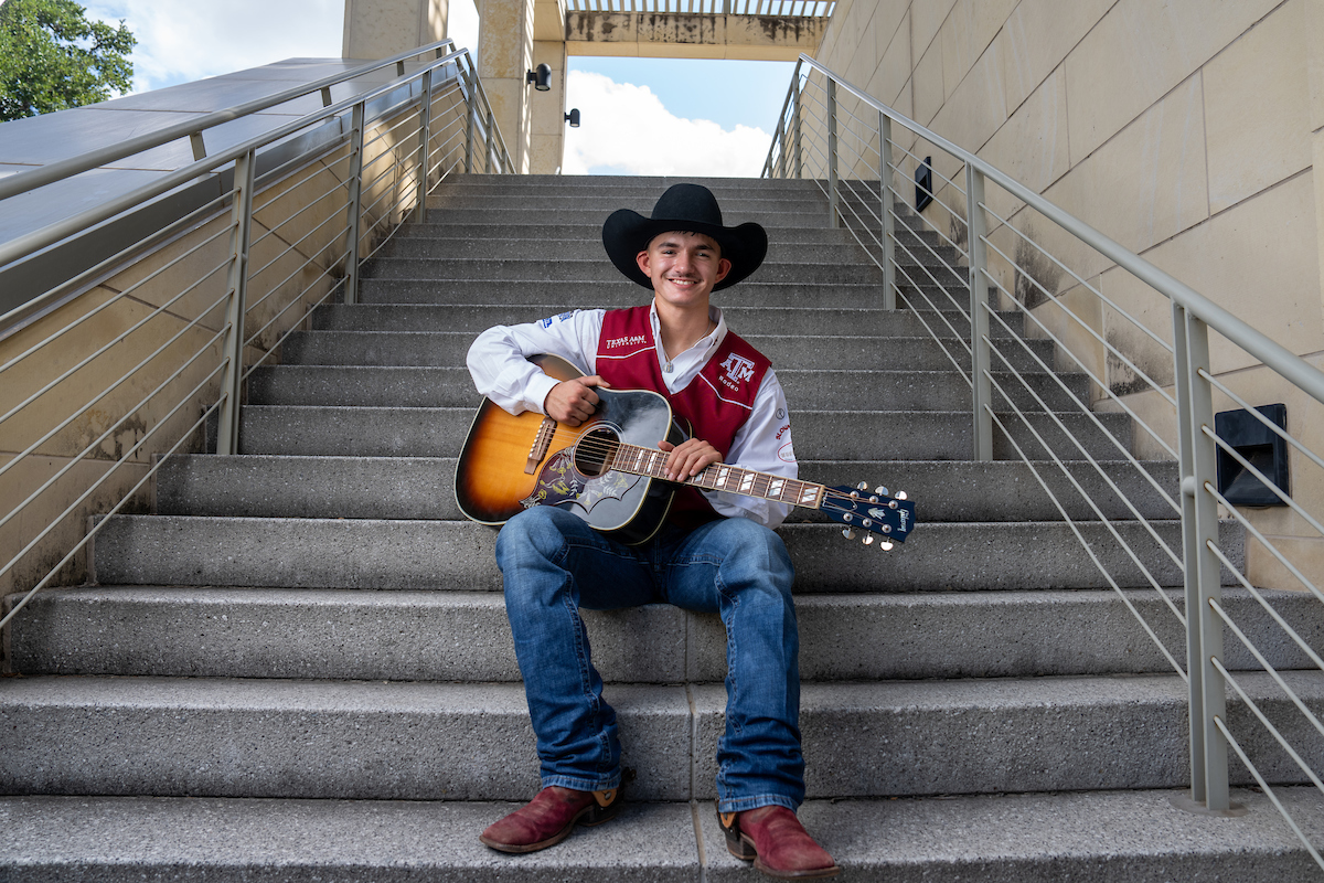 A man wearing a cowboy hat and jeans is seated on a set of stairs while holding an acoustic guitar