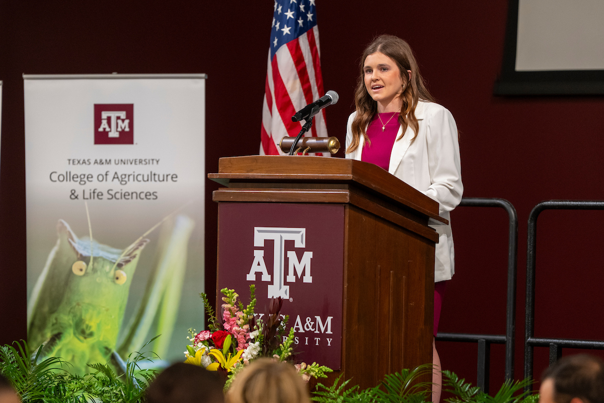 A young woman, Brittley Bowers, wearing a pink dress with a white sweater speaks from behind a podium with the Texas A&M logo.